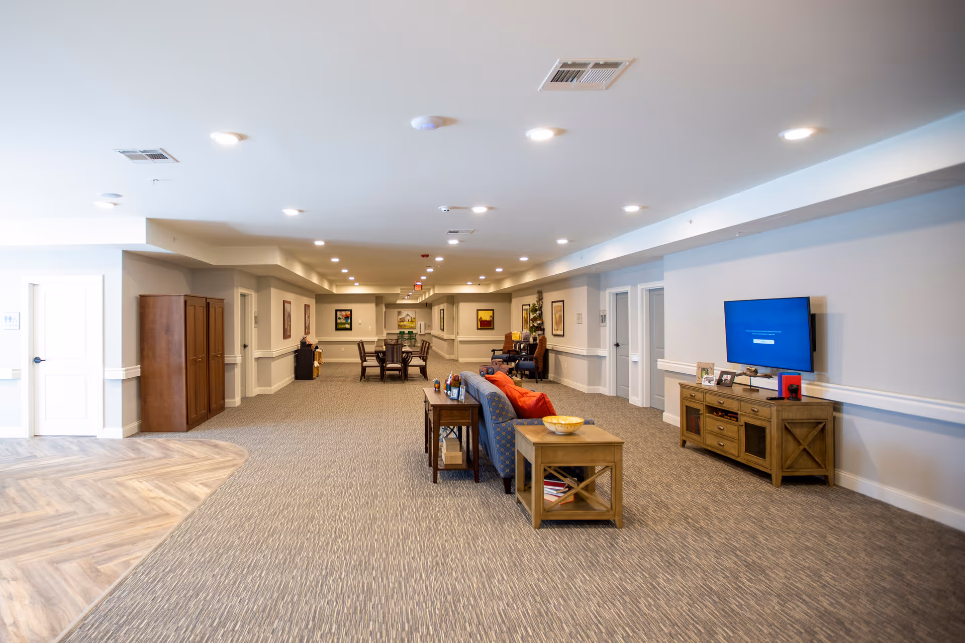 Long carpeted communal lounge and hallway with sofas, tables, a dining area in the distance, and a wall-mounted TV.
