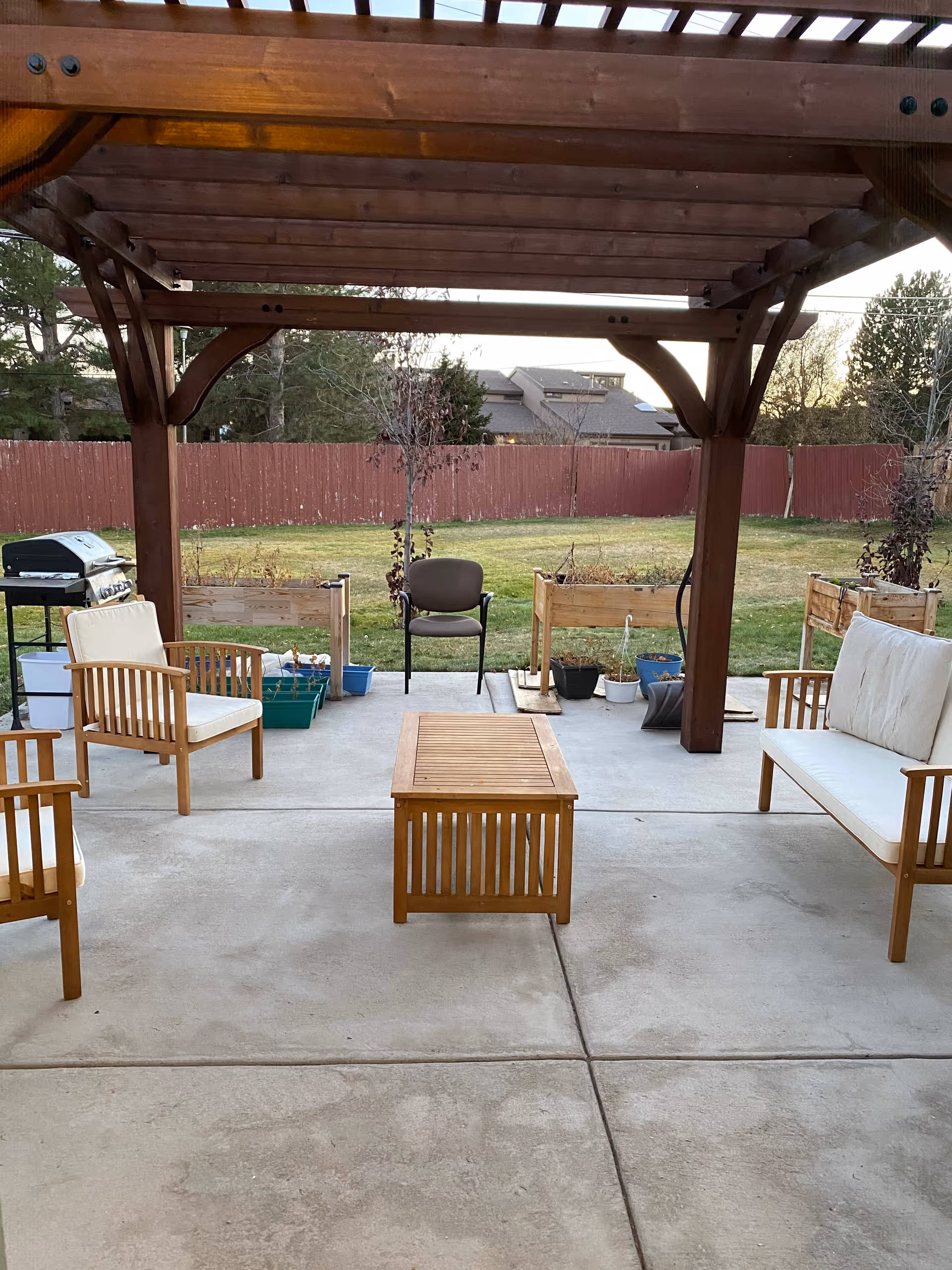 Outdoor patio area with a wooden pergola overhead, featuring cushioned wooden chairs and a wooden coffee table on a concrete floor. There are raised garden beds with plants and a barbecue grill on the left side. A red wooden fence and trees are visible in the background.