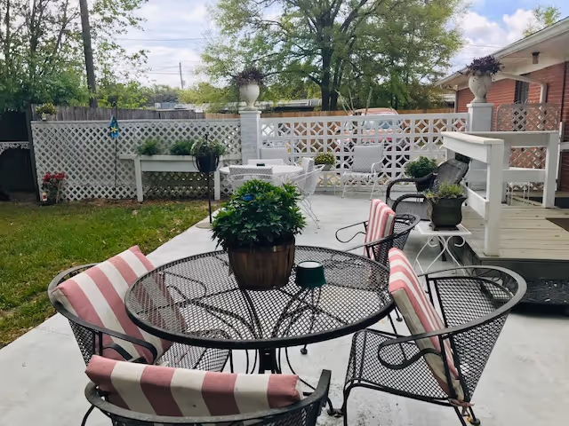 Outdoor patio area with a round metal table and four metal chairs with pink and white striped cushions. A potted plant sits on the table. In the background, there is a white lattice fence, additional seating, and greenery including trees and grass.