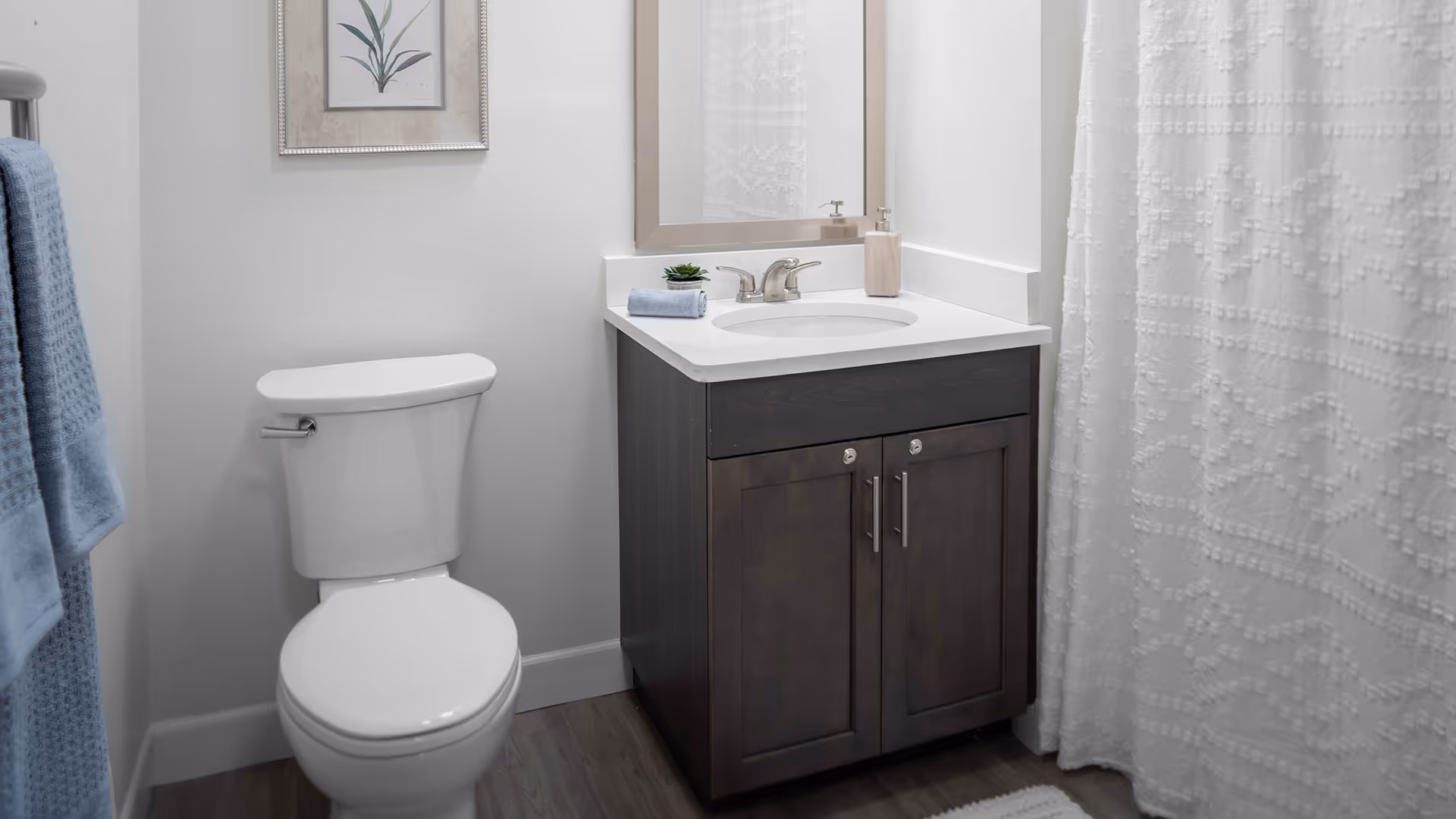 A clean and modern bathroom featuring a white toilet, a dark wood vanity with a white countertop and sink, a rectangular mirror above the sink, a small potted plant and a soap dispenser on the countertop, a blue towel hanging on a towel rack, a framed botanical artwork on the wall, and a white textured shower curtain.