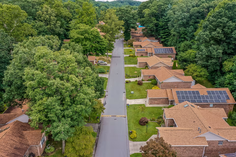 Aerial view of a senior living community street lined with single-story houses featuring brown roofs, some with solar panels, surrounded by green lawns and large trees.
