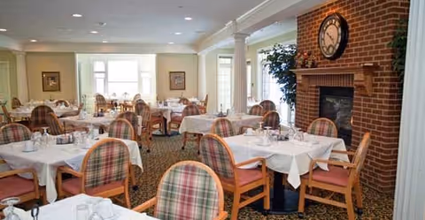 A spacious dining room with multiple tables covered in white tablecloths, each set with glassware and silverware. The room features wooden chairs with plaid cushions, a brick fireplace with a large clock above it, and large windows allowing natural light to fill the space.