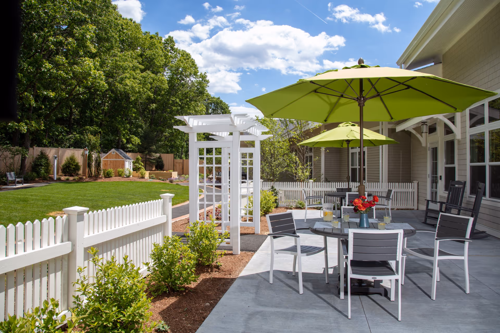 Outdoor patio area with tables and chairs under large green umbrellas next to a building. The patio is surrounded by a white picket fence and overlooks a well-maintained lawn with trees and a small shed in the background under a partly cloudy sky.