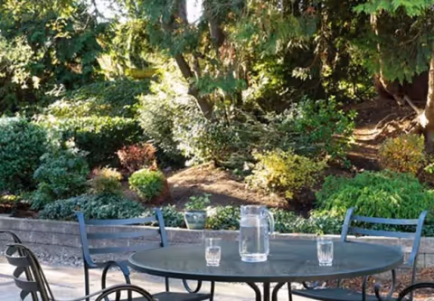Outdoor patio area with a round metal table and four metal chairs. On the table, there is a clear glass pitcher filled with water and three empty glasses. The background features lush green bushes, trees, and a stone retaining wall.