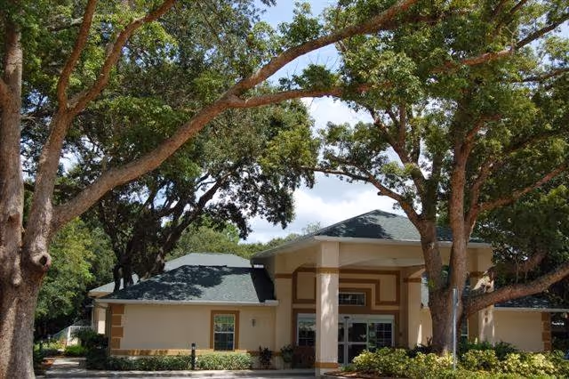 Exterior view of Accordia Woods Assisted Living Community building surrounded by large trees and greenery under a partly cloudy sky.