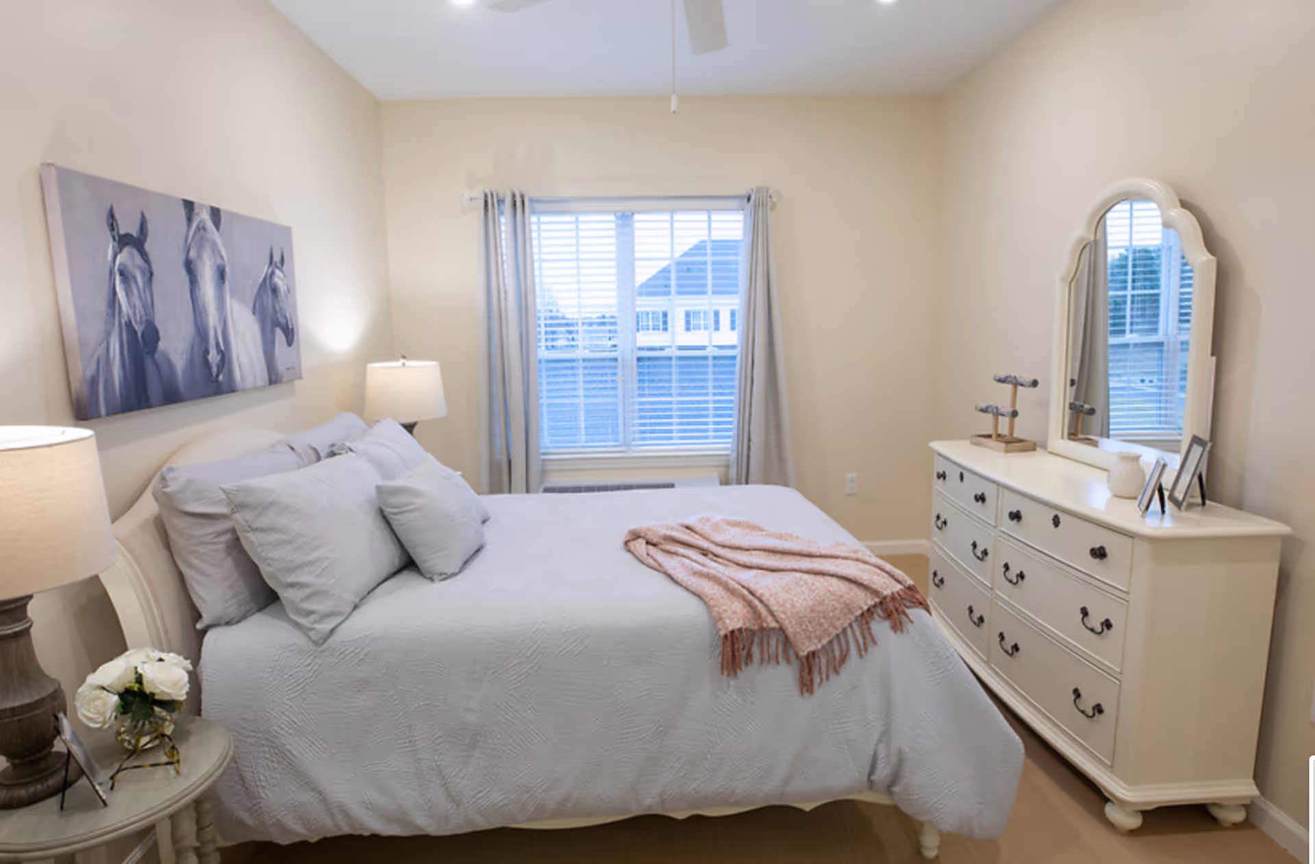 A cozy bedroom with a neatly made bed featuring light gray bedding and a pink throw blanket. There are two bedside tables with lamps and a small vase of white flowers on one side. A large window with light curtains lets in natural light. On the right side, there is a white dresser with a mirror and decorative items on top. A painting of three horses hangs above the bed.