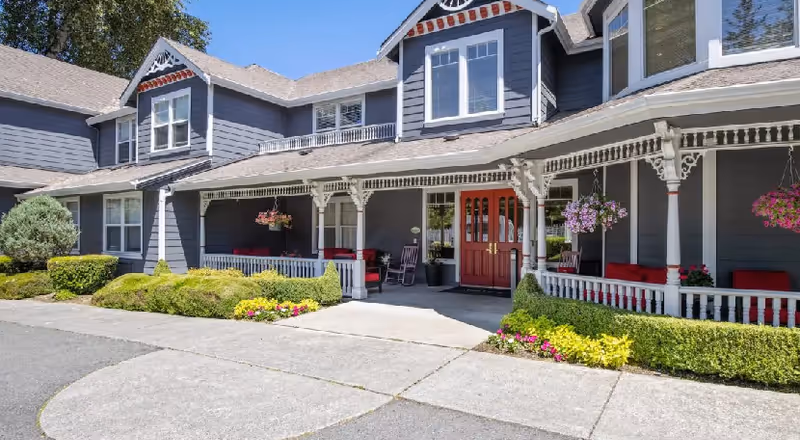Exterior view of a senior living facility named Aegis Lodge featuring a large porch with white decorative railings and columns, hanging flower baskets, red cushioned seating, and a red double door entrance. The building is painted gray with white trim and surrounded by neatly trimmed bushes and colorful flowers.