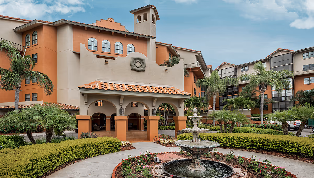 Exterior view of Lake Seminole Square, featuring a multi-story building with terracotta roof tiles and arched architectural details. The foreground includes a landscaped garden with palm trees, shrubs, and a tiered water fountain surrounded by a circular walkway.