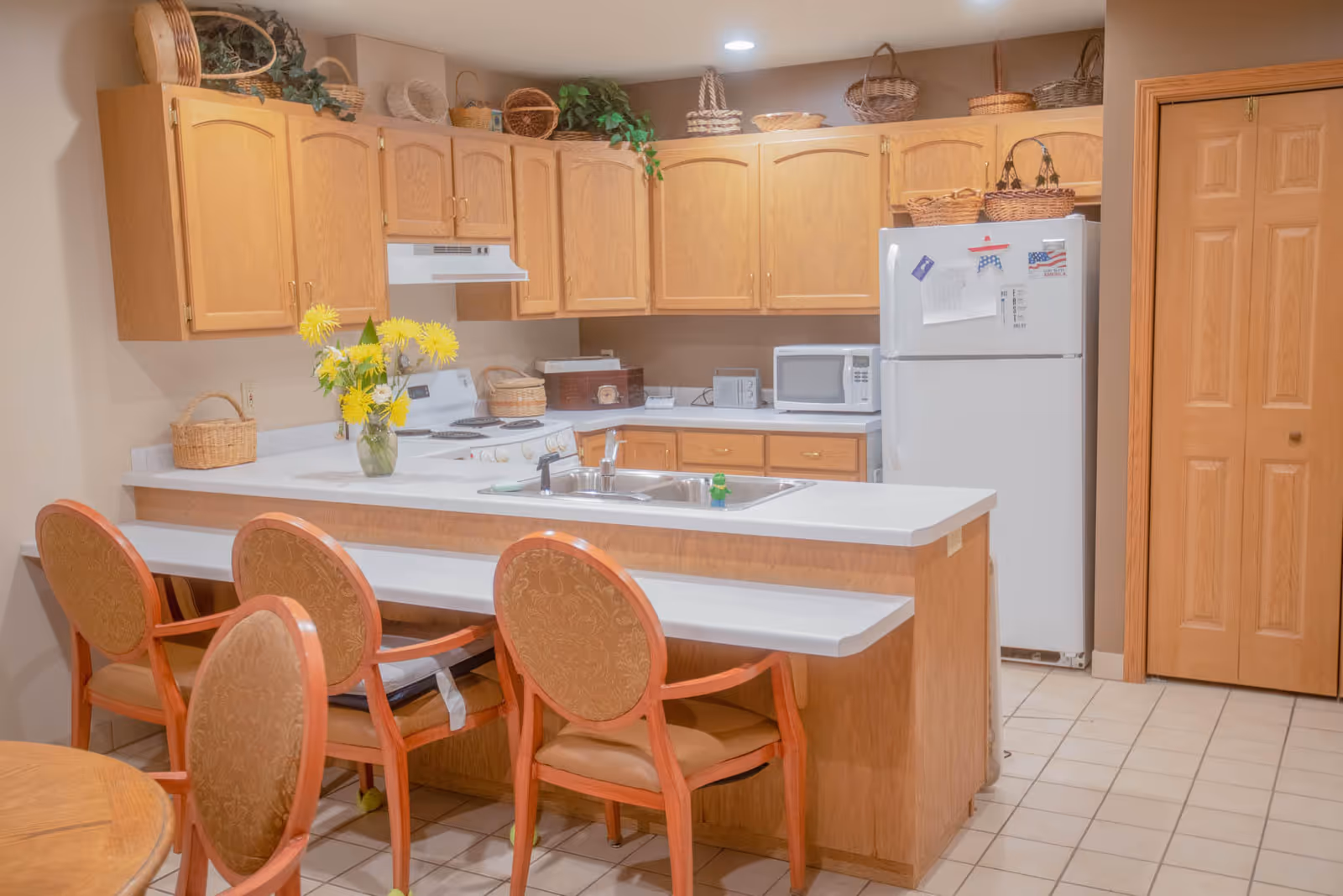 A kitchen area with wooden cabinets, a white refrigerator, microwave, stove, and sink. There are several wicker baskets on top of the cabinets and a vase with yellow flowers on the counter. Four wooden chairs with cushioned seats are placed around a counter and a small table. The floor is tiled and there is a wooden door to the right.