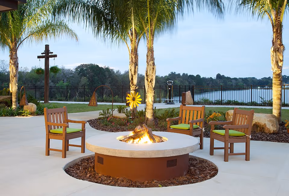 Outdoor patio area with a circular fire pit surrounded by four wooden chairs with green cushions. Palm trees and decorative garden elements are visible, with a lake and trees in the background under a clear sky.