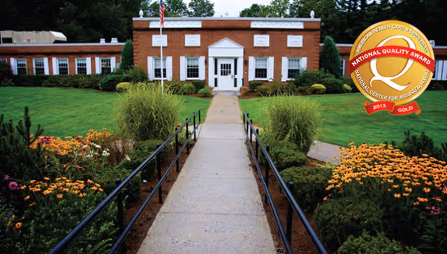 Front entrance of a red-brick nursing care building with a paved walkway, lawns and flowerbeds, and a quality award badge in the top-right.