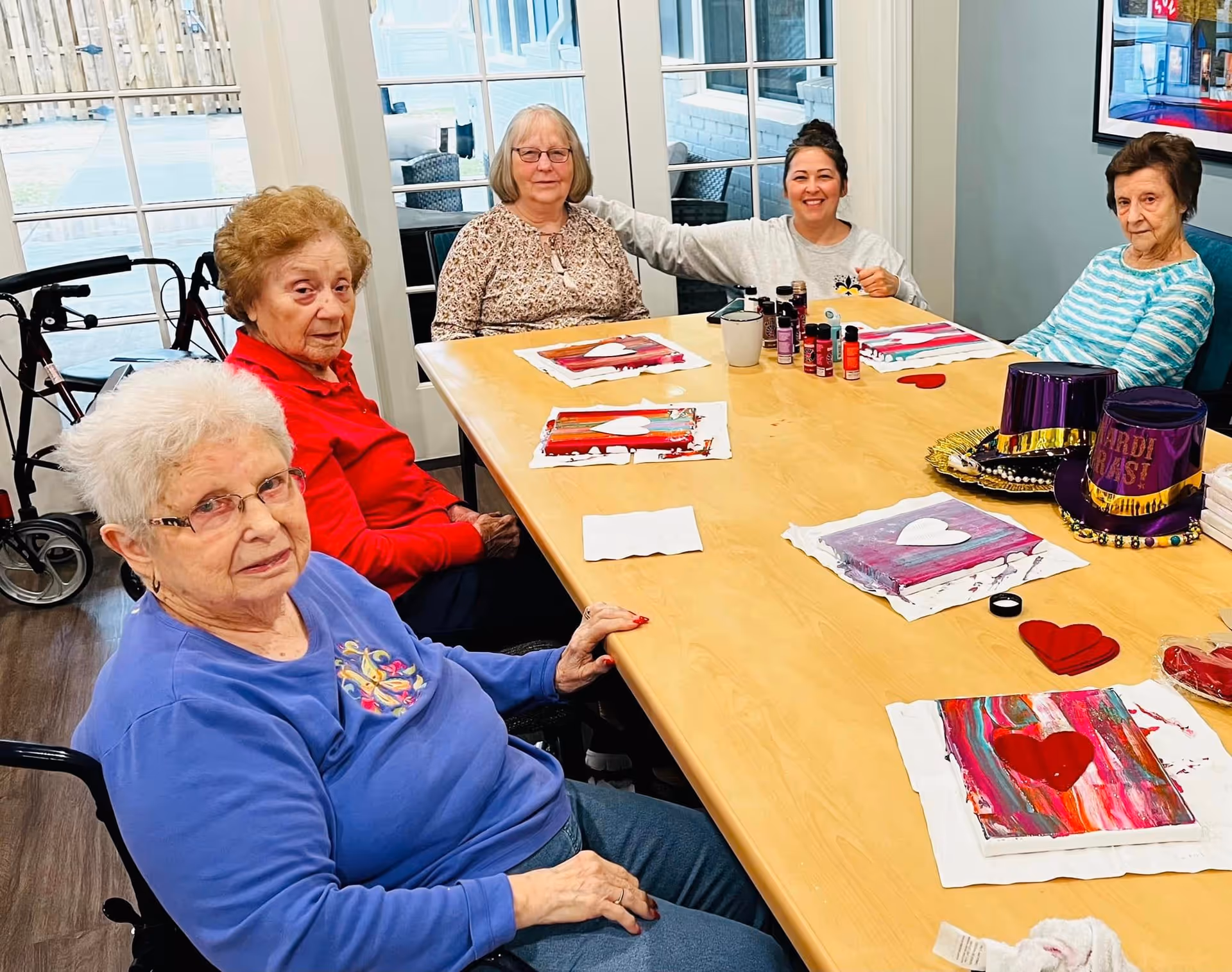 Five elderly women sitting around a wooden table in a bright room with large windows. They appear to be engaged in a painting activity with colorful canvases featuring heart shapes and various paint bottles on the table. One woman is smiling and has her arm around another. There are two purple Mardi Gras hats on the table.