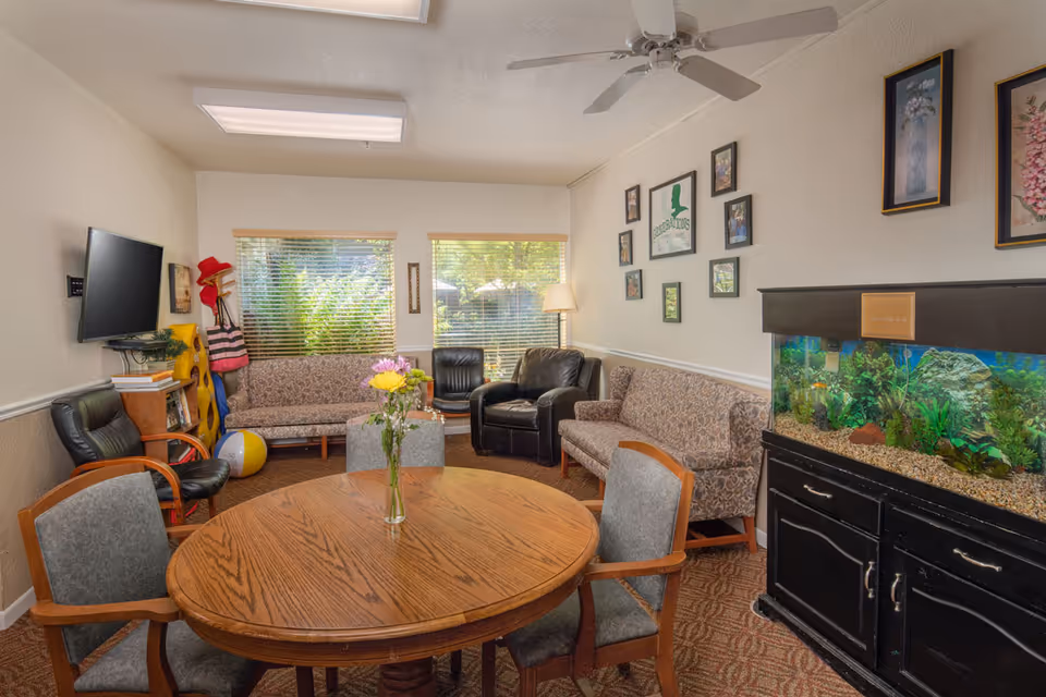 A cozy living room with a round wooden table and four chairs in the foreground. The room has two patterned sofas, two black armchairs, a large fish tank on a black cabinet, and a wall-mounted TV. There are framed pictures and artwork on the walls, a ceiling fan, and two windows with blinds letting in natural light. A vase with flowers is on the table.