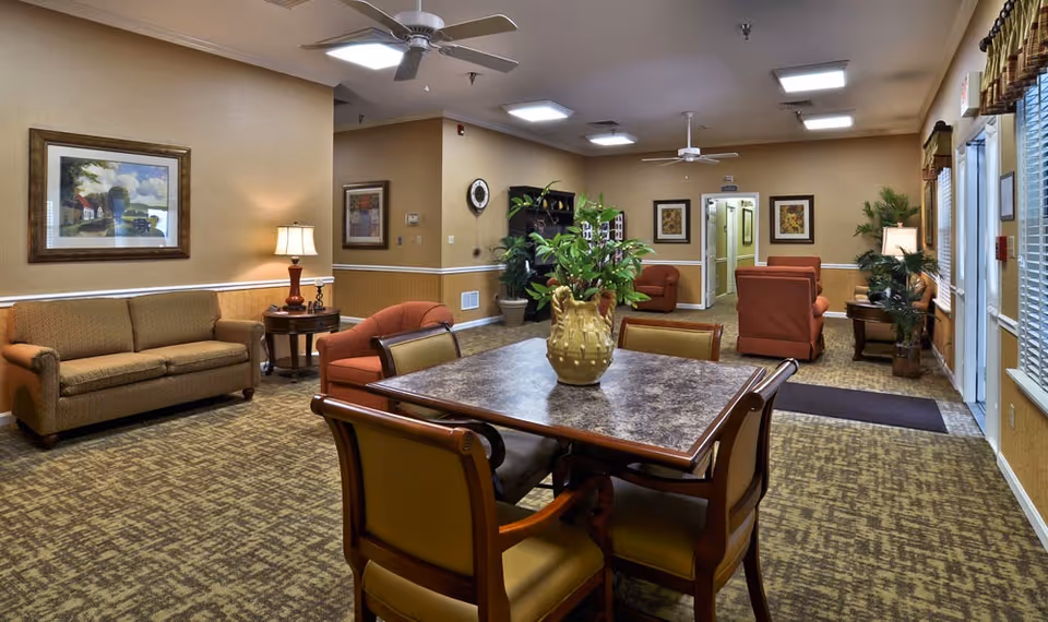 A cozy living room area in a senior living facility with a square table and four chairs in the foreground, a large plant centerpiece on the table, multiple armchairs and a sofa along the walls, framed artwork, table lamps, ceiling fans, and large windows with blinds and valances.