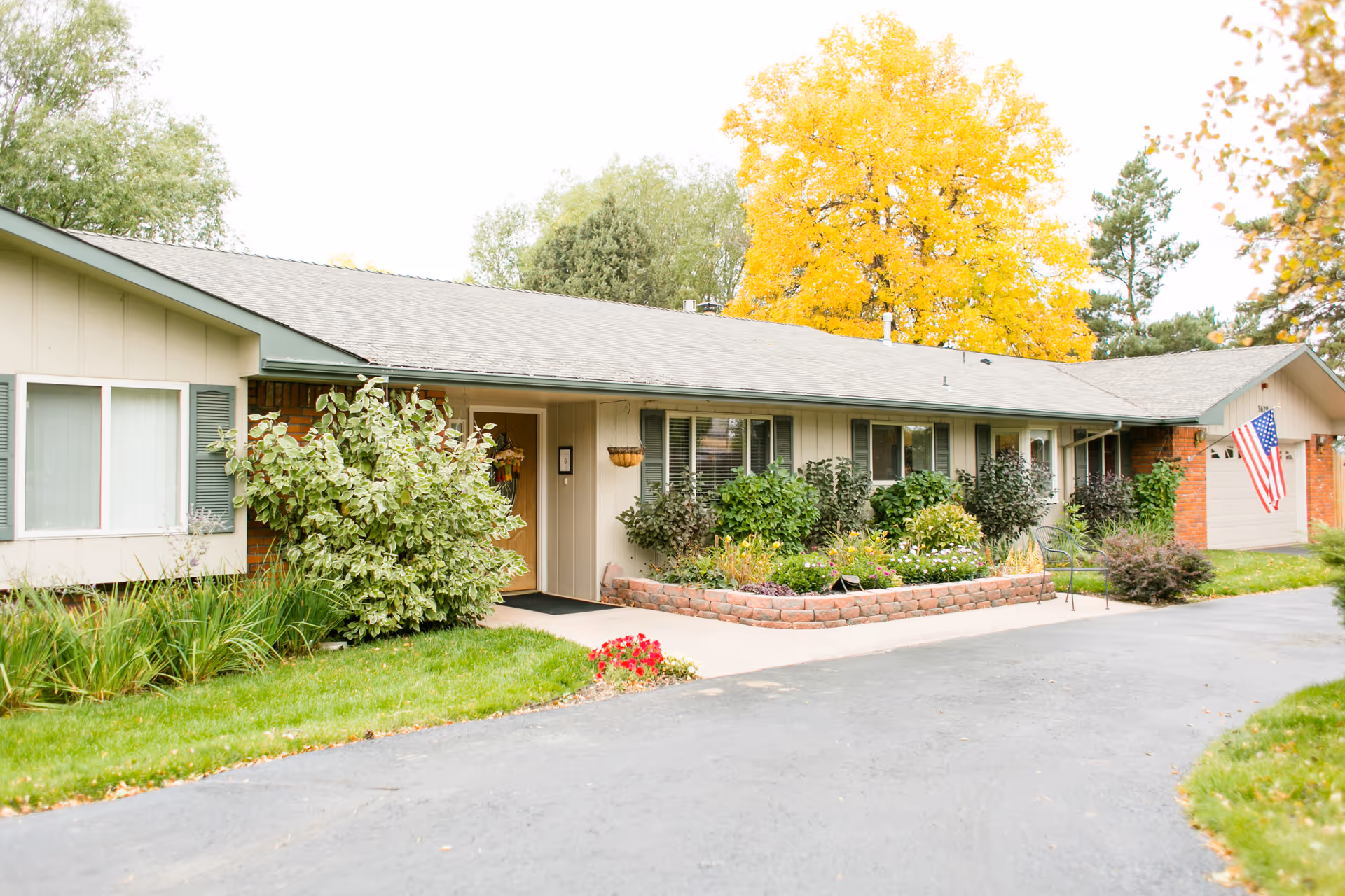 Single-story assisted living building with a landscaped front garden, driveway, and an American flag.