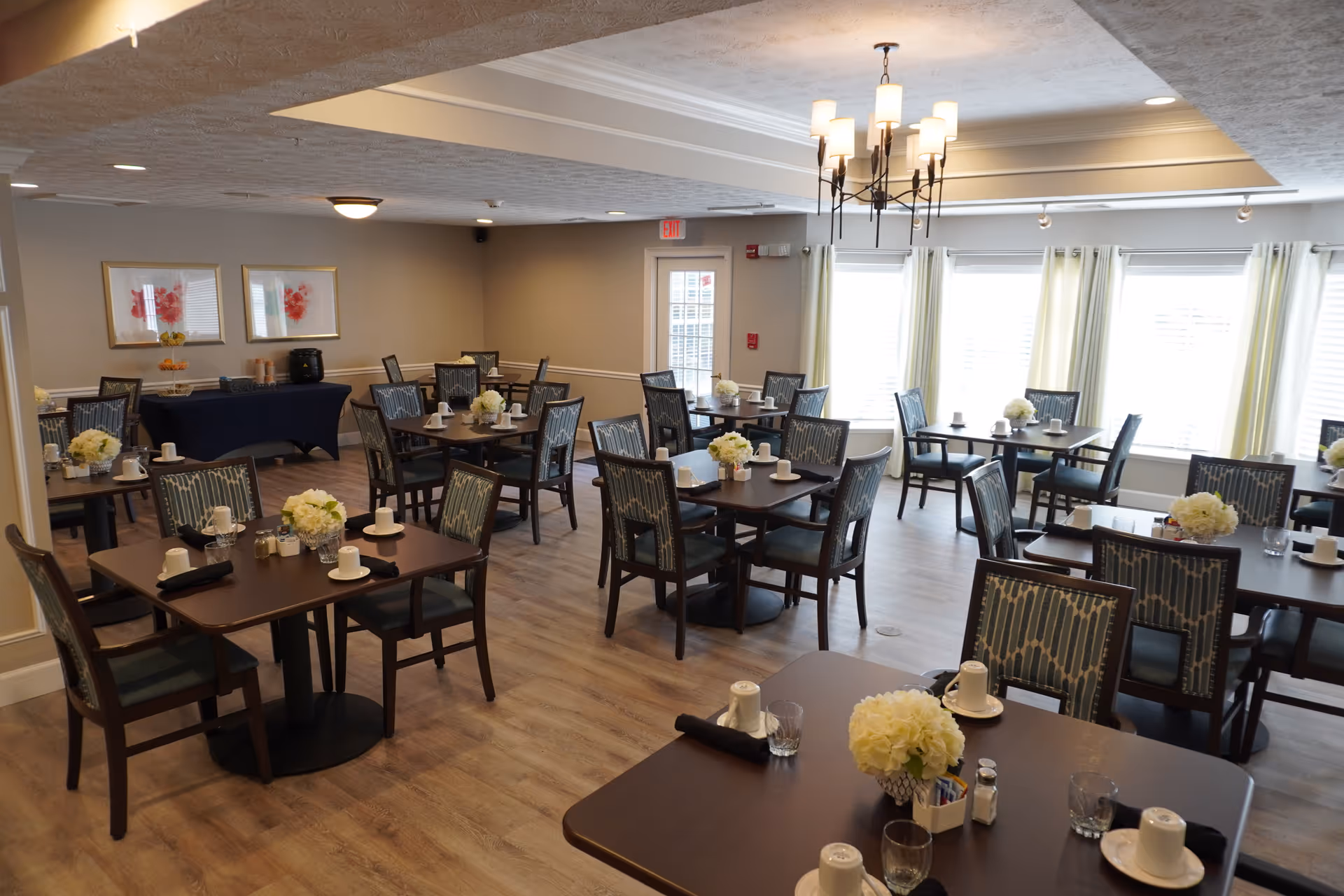 A bright dining room with multiple dark wood tables and chairs arranged neatly. Each table is set with white cups, glasses, black napkins, and small flower arrangements. The room has large windows with light curtains allowing natural light to fill the space. A chandelier hangs from the ceiling, and two framed floral artworks are visible on one wall.