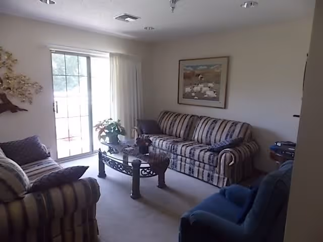 A living room with a striped sofa and loveseat, a blue armchair, a glass coffee table with decorative items, a large window with sliding glass door and curtains, a wall painting, and a small side table with a lamp.