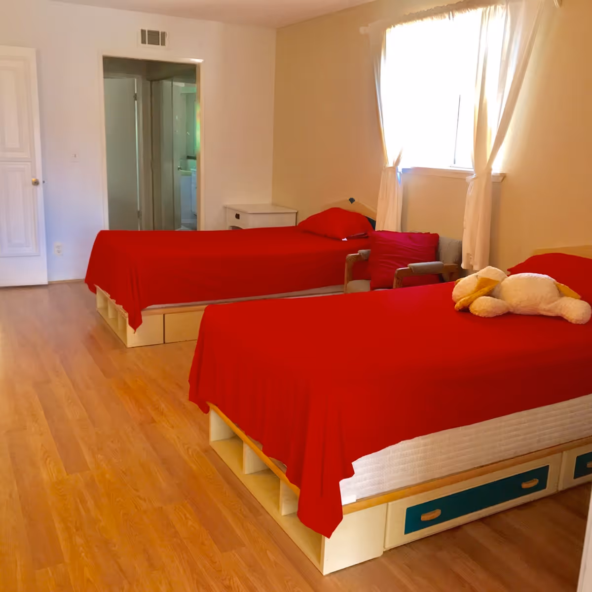 A bedroom with two single beds covered in red bedspreads. There is a small white nightstand between the beds, a window with white curtains letting in natural light, and a stuffed animal on one of the beds. The room has wooden flooring and light-colored walls.