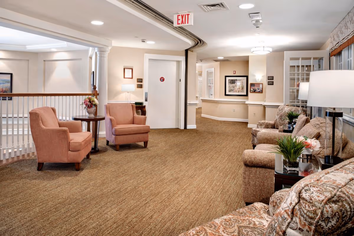 A cozy senior living facility common area with beige walls and carpet. The space features two pink upholstered armchairs with a small round table and flower arrangement between them on the left. On the right, there are patterned sofas and armchairs with side tables holding lamps and small plants. The area is well-lit with ceiling lights and has framed artwork on the walls. An exit sign is visible above a white door in the background.