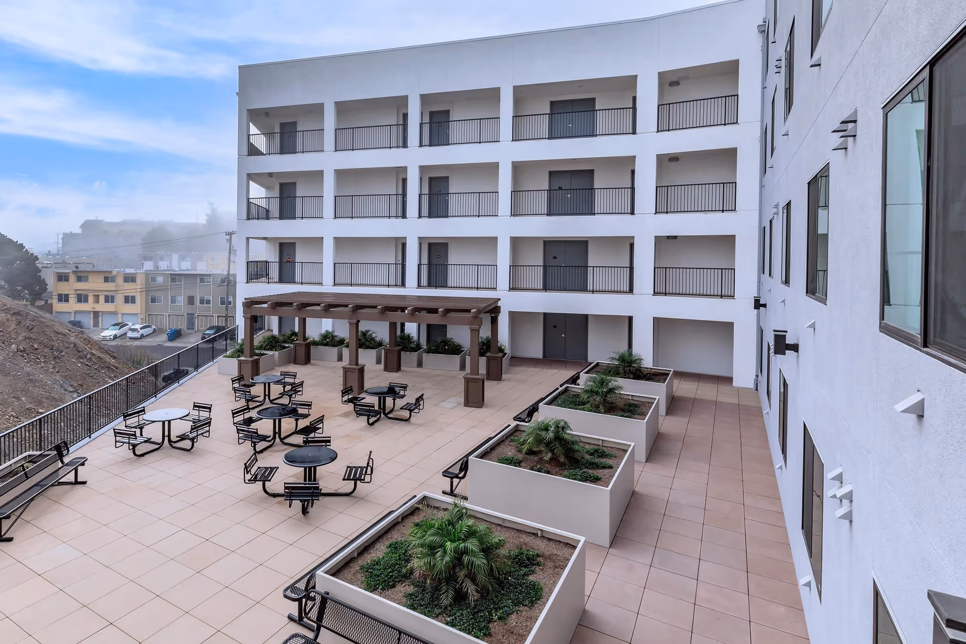 Outdoor courtyard area of Brunswick Street Apartments featuring multiple round tables with attached seating, benches, large planter boxes with greenery, and a wooden pergola. The courtyard is surrounded by a multi-story white building with balconies and doors overlooking the space.
