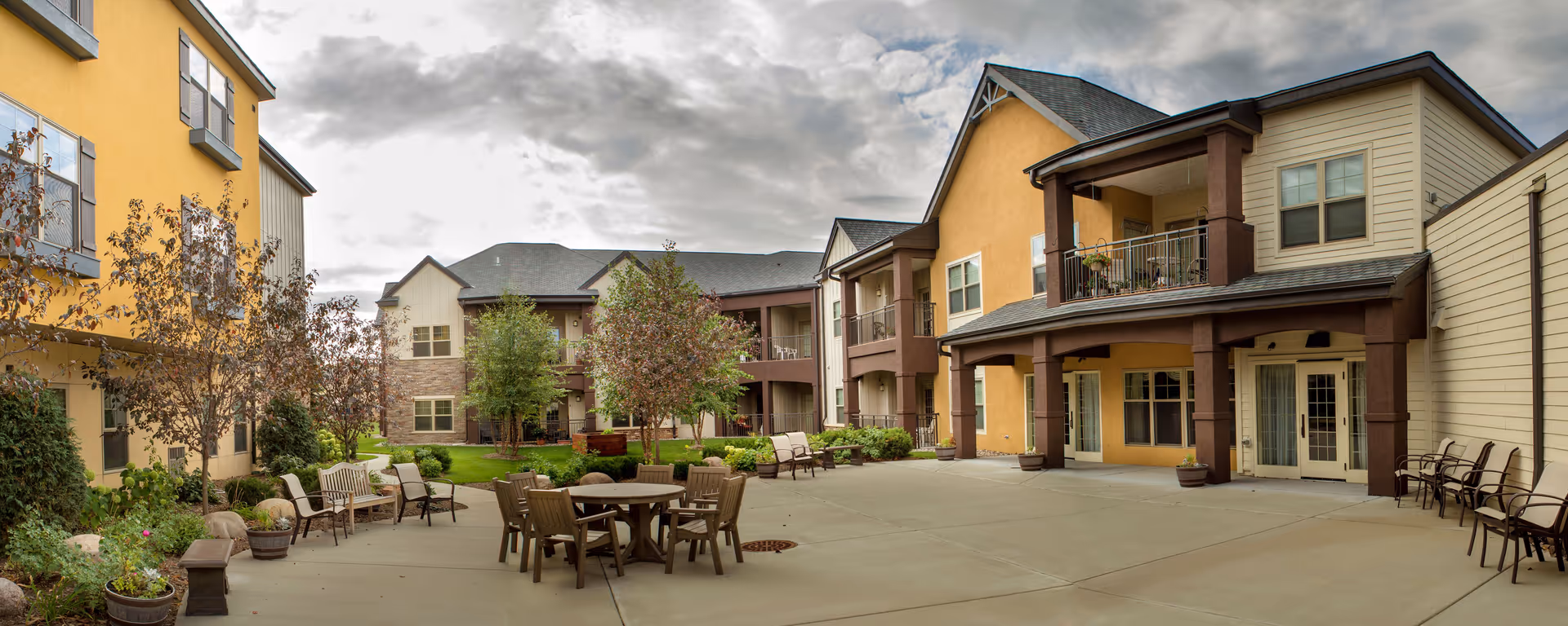 Sunny courtyard with patio tables, chairs, planters and landscaped trees surrounded by a multi-story senior living building with balconies.