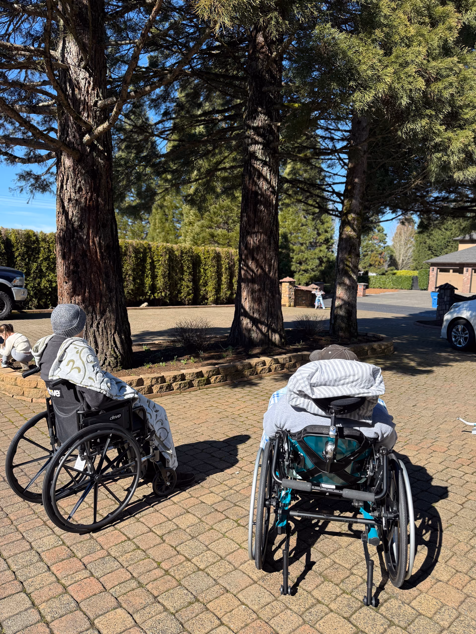 Two elderly individuals in wheelchairs sitting outside on a paved area under tall trees, enjoying the sunny day at Sequoia Meadows Senior Care. One person is covered with a blanket and wearing a hat, while the other is also covered with a blanket. There are cars and greenery in the background.