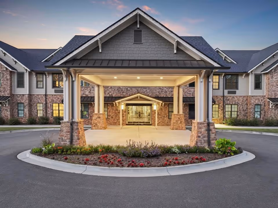 Front exterior view of a senior living facility named Brickmont Roswell at dusk, featuring a covered entrance with stone pillars, a landscaped roundabout with flowers, and a brick and siding building with multiple windows.