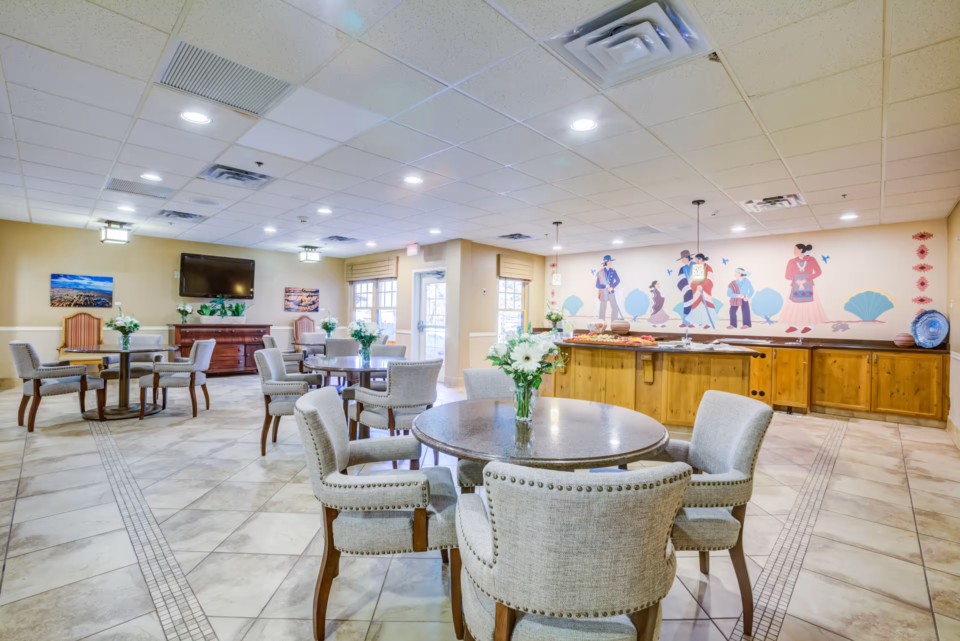 A bright and spacious dining area in a senior living community featuring round tables with upholstered chairs, floral centerpieces, a wooden buffet counter with food trays, and a colorful mural of people in traditional attire on the wall. The room has tiled floors, recessed lighting, and large windows allowing natural light.