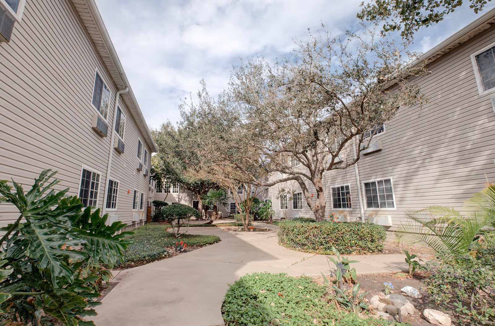 Outdoor courtyard area at Garden Estates of Corpus Christi featuring a paved walkway surrounded by green shrubs, trees, and plants, with beige two-story buildings on either side under a partly cloudy sky.