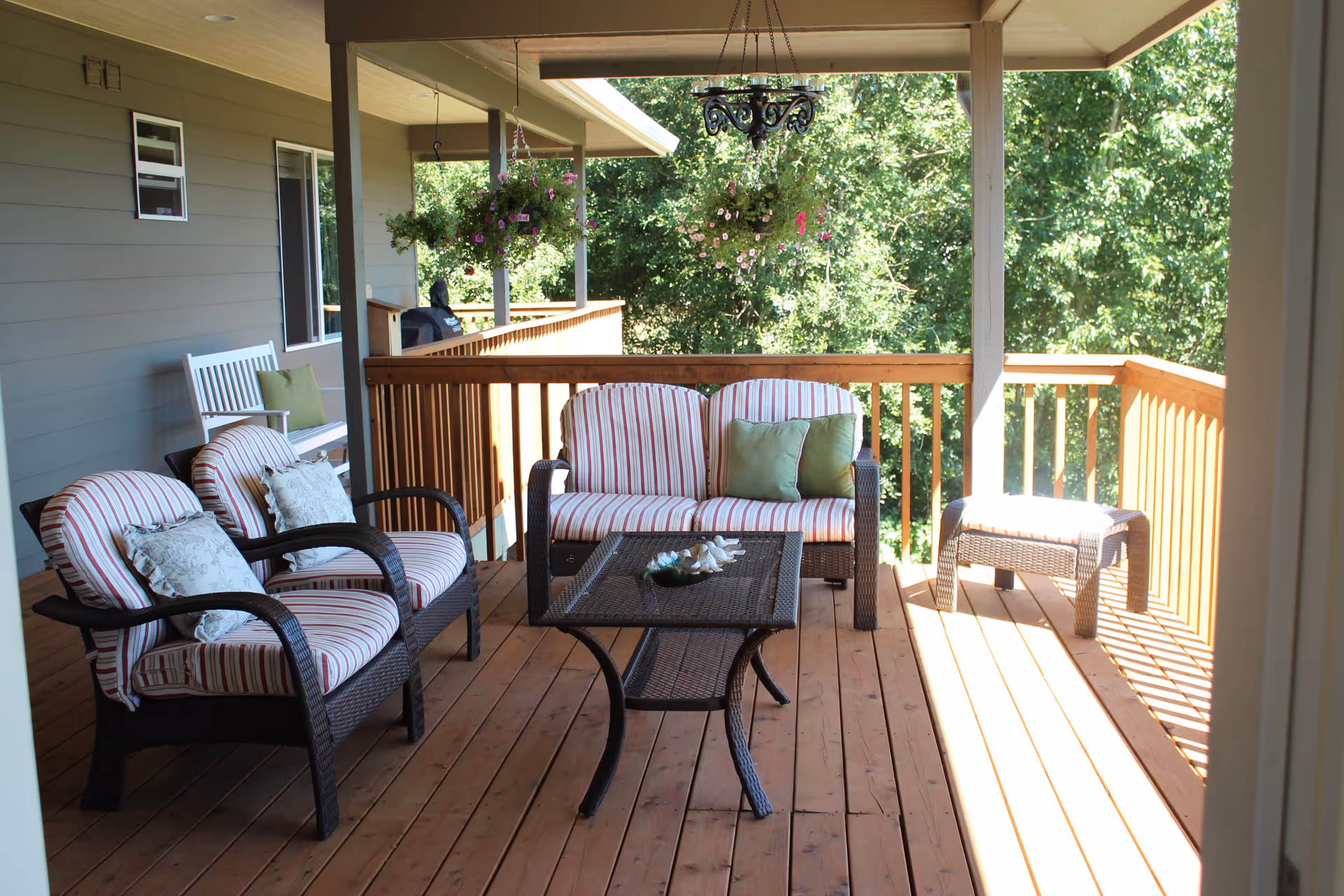 A wooden outdoor porch with cushioned wicker furniture including two armchairs, a loveseat, a coffee table, and a small side table. There are hanging flower baskets and a white bench with green cushions against the house wall. Trees and greenery are visible in the background.