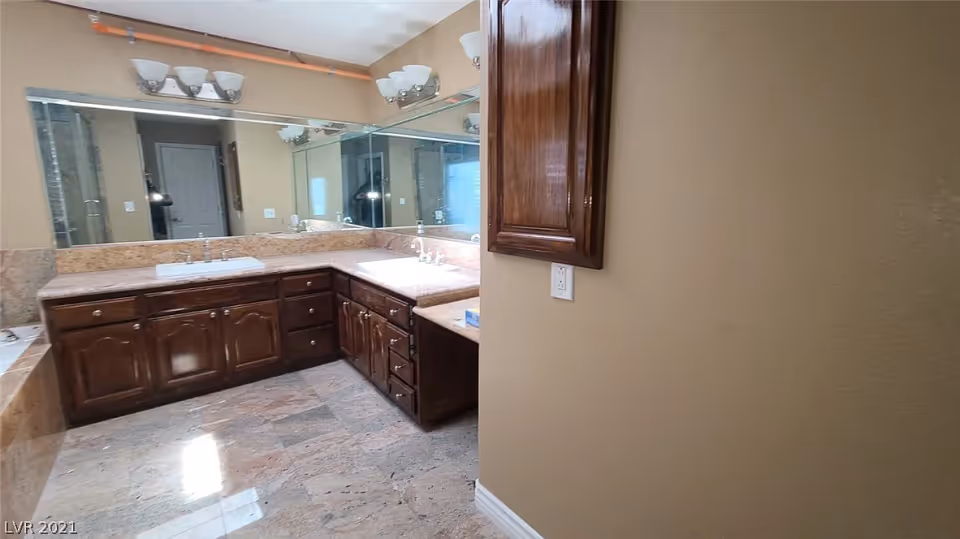 A spacious bathroom with a large corner vanity featuring two sinks, dark wooden cabinets, and a wide mirror above. The floor and countertop are made of matching stone tiles, and there are two light fixtures with three bulbs each mounted above the mirror. A wooden cabinet is mounted on the beige wall to the right.