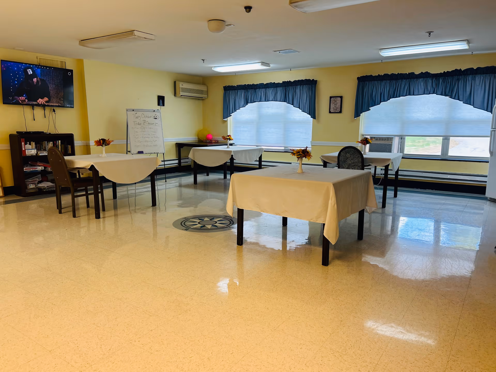 A bright room with four tables covered with white tablecloths, each with a small flower arrangement. The room has large windows with blue curtains, a TV mounted on the wall showing a person, a whiteboard with writing, and a bookshelf. The floor is shiny and light-colored.