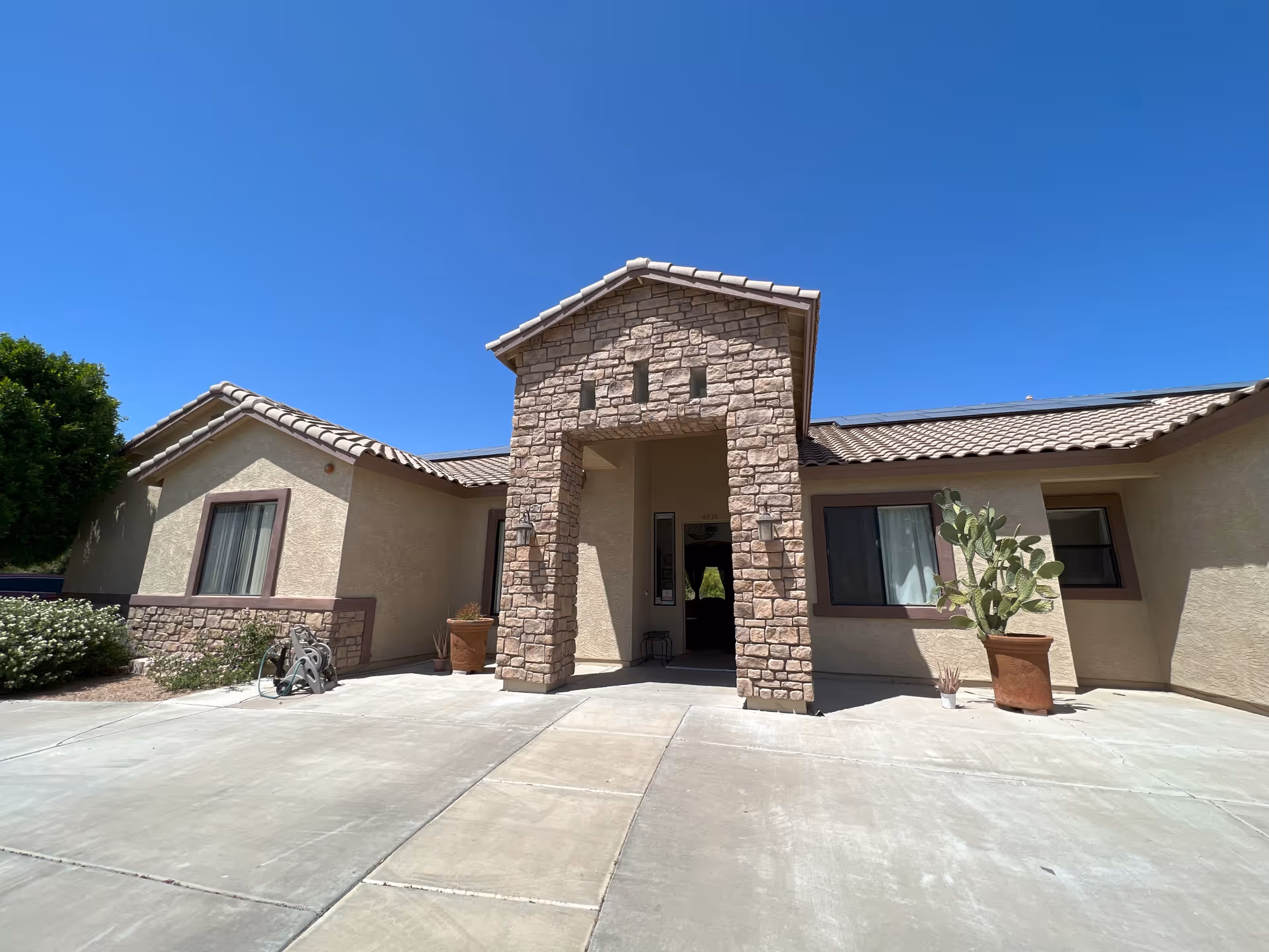 Front exterior view of a single-story building with beige stucco walls and a stone archway entrance under a clear blue sky. There are potted plants and a small garden area near the entrance.