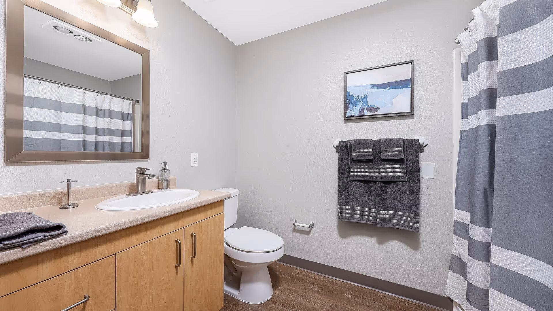 A clean and modern bathroom featuring a wooden vanity with a white sink and silver faucet, a large rectangular mirror above the sink, a toilet next to the vanity, and a shower with a gray and white striped curtain. On the wall above the toilet, there is a towel rack holding neatly folded dark gray towels and a framed abstract painting. The floor is wood-style, and the walls are painted light gray.