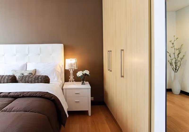 A modern bedroom featuring a white tufted headboard bed with brown and white bedding, a white nightstand with a clear lamp and a small vase of white flowers, light wood wardrobe with silver handles, and a large mirror reflecting a vase with branches in the adjacent room.
