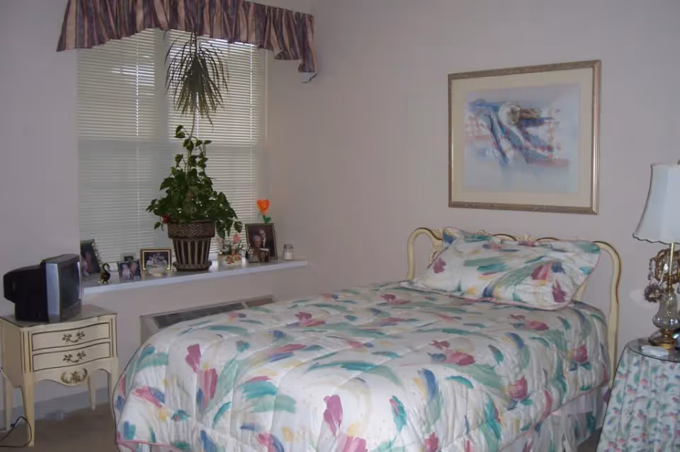 A bedroom with a single bed covered in a floral patterned comforter and matching pillow. There is a small vintage nightstand with a small TV on it to the left of the bed. A window with closed blinds and a valance is behind the bed, with a windowsill holding several framed photos and a potted plant. On the right side of the bed is a small round table with a floral tablecloth and a decorative lamp. A framed abstract painting hangs on the wall above the bed.