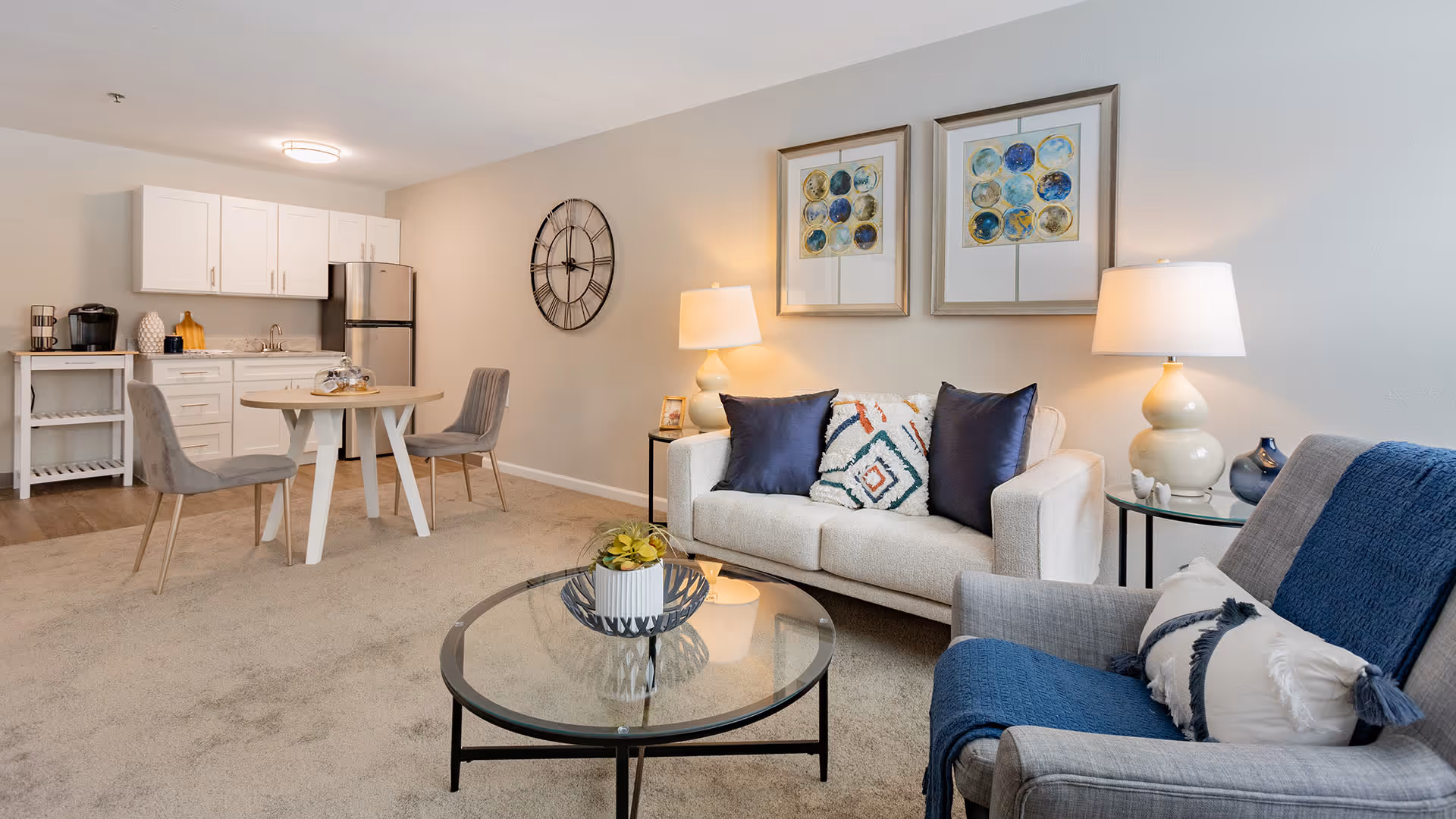 A cozy living room and kitchen area in a senior living facility. The living room features a white sofa with decorative pillows, a gray armchair with a blue throw blanket, a glass coffee table with a small plant, and two table lamps on side tables. The kitchen area has white cabinets, a stainless steel refrigerator, a small round dining table with two gray chairs, and a coffee maker on a small white shelf. The walls are decorated with two framed abstract art pieces and a large wall clock.