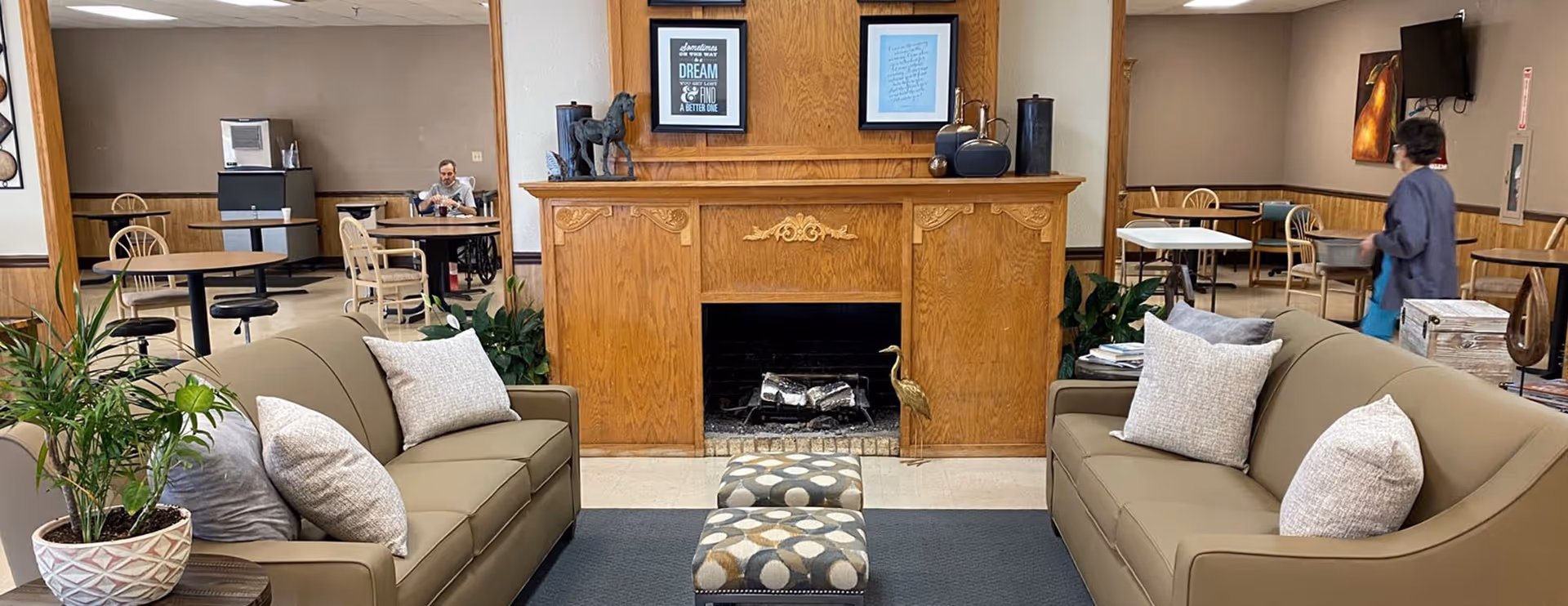 A cozy common area in a senior living facility with two beige sofas facing each other, a wooden fireplace with decorative items on the mantel, two patterned ottomans in front of the fireplace, and several tables and chairs in the background. There are plants on either side of the fireplace and a person sitting at a table in the back left, while another person is walking on the right side of the room.