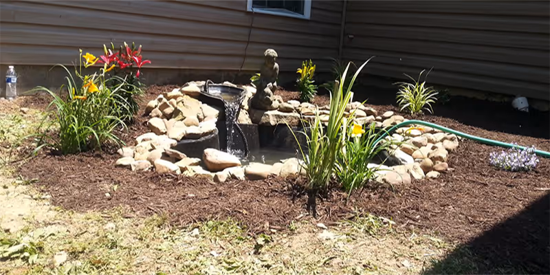 A small outdoor garden area featuring a decorative rock water fountain with water flowing into a small pond. The garden is surrounded by mulch and has various flowering plants including yellow and red flowers. A green garden hose is visible on the right side near the building's exterior wall.