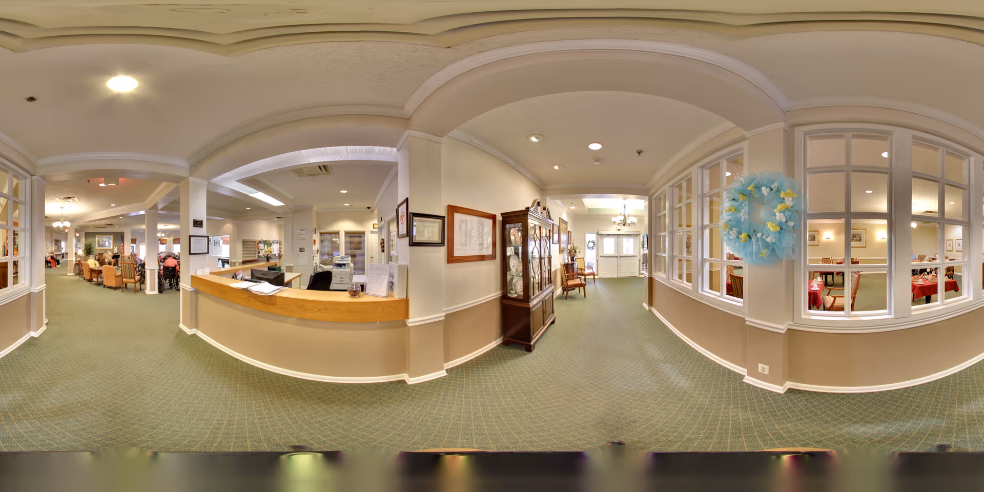 Interior lobby and reception area of a senior living facility with a front desk, seating area, hallway, and dining room visible through windows.