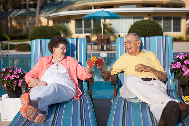 An elderly man and woman sitting on striped lounge chairs by a pool, smiling and clinking their glasses filled with red beverages garnished with orange slices. There are potted pink flowers on either side of the chairs and a building with windows and a patio umbrella in the background.