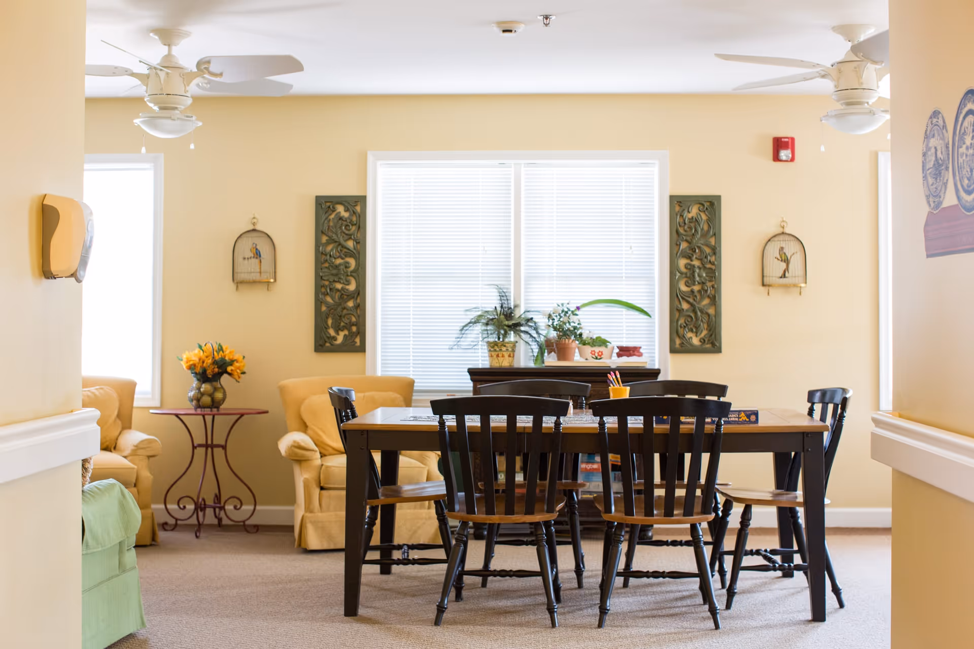 A cozy common area with a wooden table surrounded by six dark wooden chairs. Behind the table are two beige armchairs with a small round table between them holding a vase of yellow flowers. The walls are light yellow with decorative wall hangings and two windows with white blinds. There are ceiling fans and some potted plants on a cabinet beneath the windows.