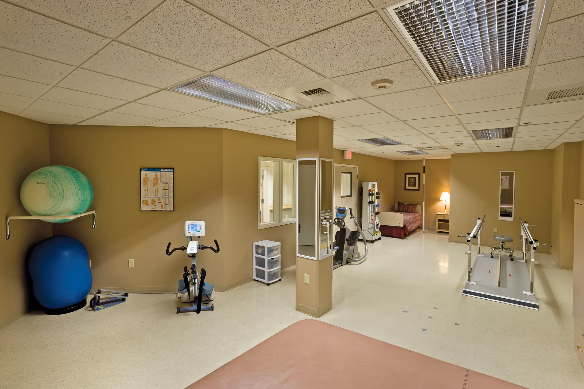 A rehabilitation or physical therapy room with exercise equipment including stationary bikes, balance balls, parallel bars for walking practice, and a small bed or treatment table. The room has beige walls, a tiled ceiling with fluorescent lights, and a small window opening to another room.