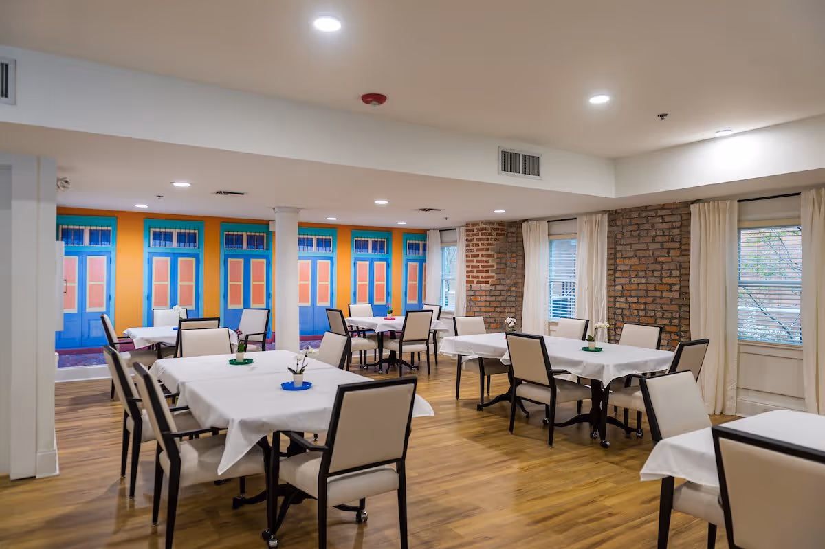 A bright dining room with several tables covered with white tablecloths and surrounded by beige cushioned chairs. The room features wooden flooring, exposed brick walls, windows with white curtains, and a colorful mural of blue and orange doors on one wall. The ceiling has recessed lighting.