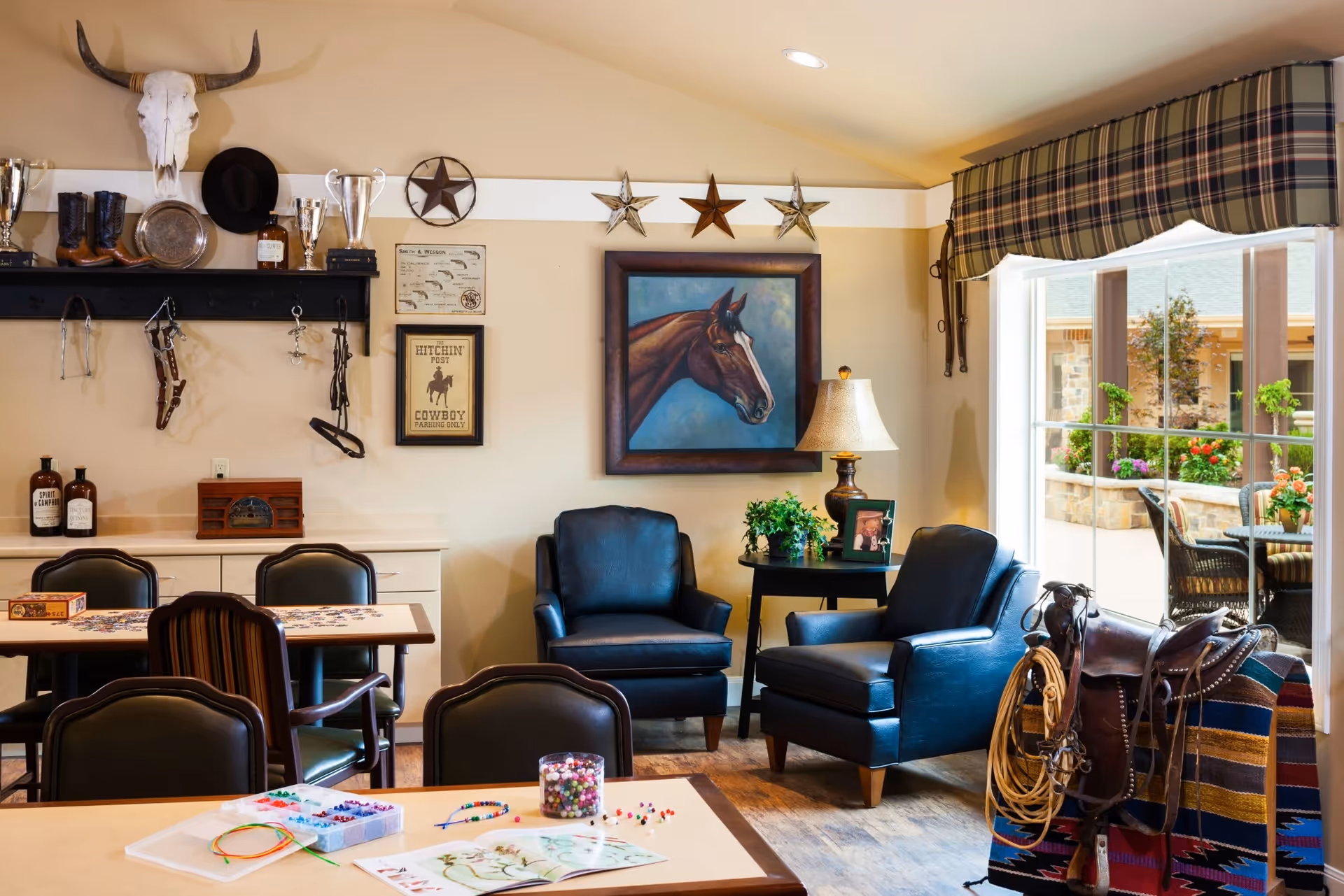 A cozy interior room with western-themed decor including a horse painting, cowboy hat, boots, and a skull mounted on the wall. The room has two dark leather armchairs, a side table with a lamp and a plant, several tables with chairs, and craft supplies on one table. A large window with plaid valance shows an outdoor patio with chairs and flowers.