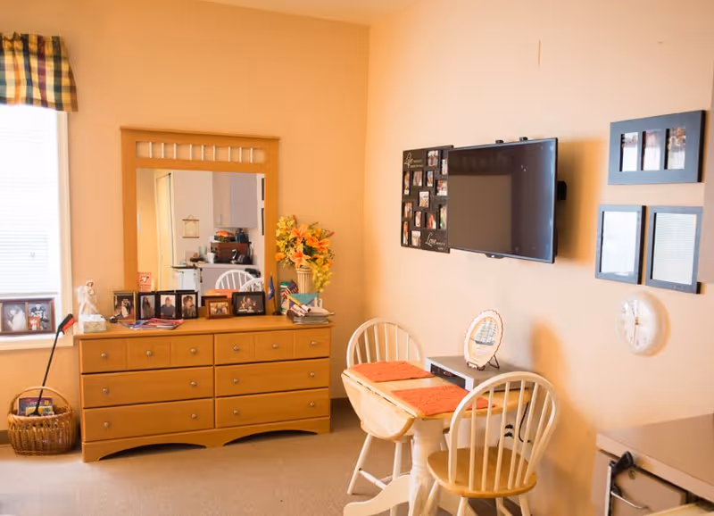 Small assisted-living room with a dresser and mirror, wall-mounted TV, and a small dining table with two chairs.