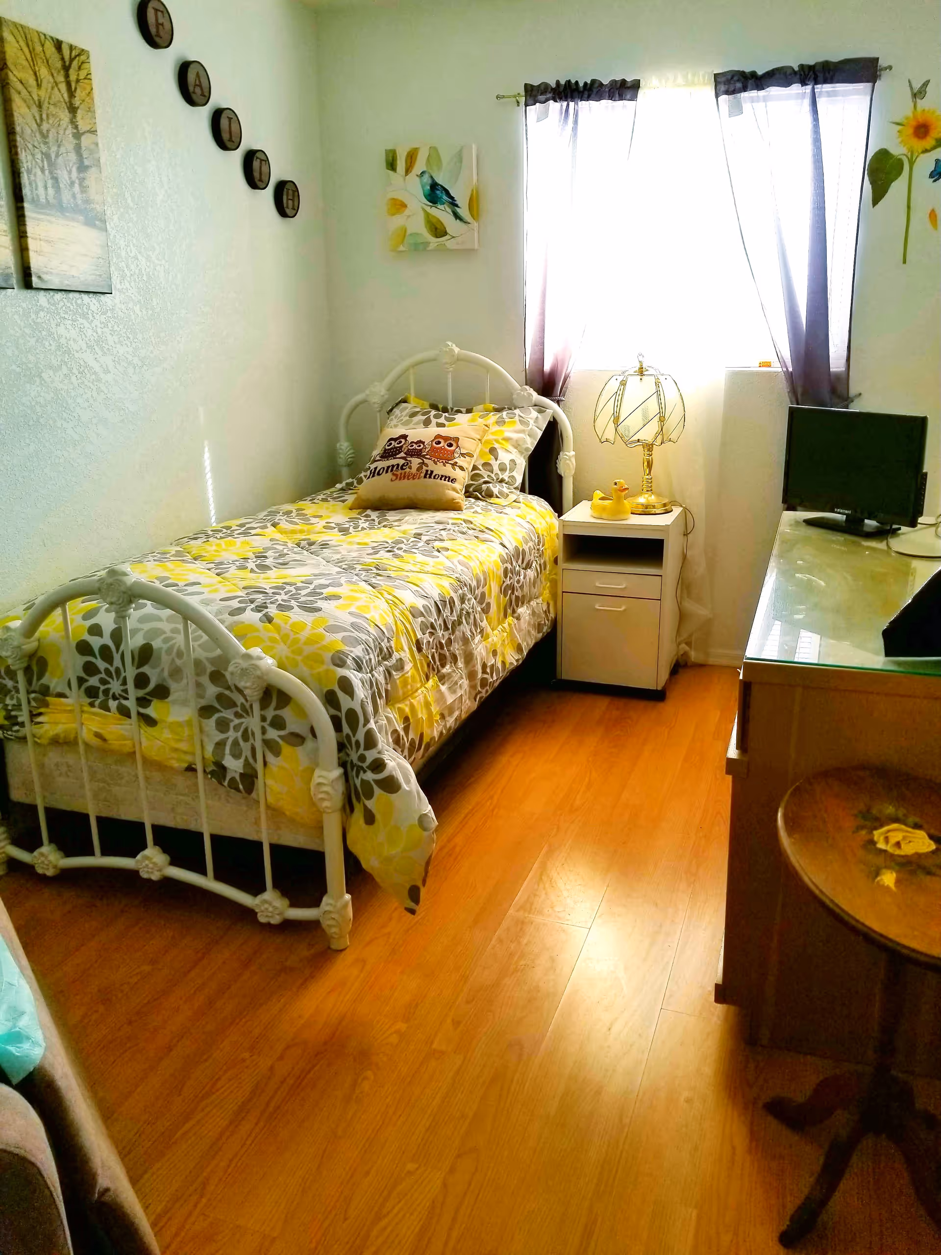A small bedroom with a single white metal bed frame, a bed covered with a yellow, gray, and white floral comforter, and a pillow with two owls and the text 'Home Sweet Home'. There is a nightstand next to the bed with a decorative lamp and two yellow rubber ducks. A window with sheer purple curtains lets in bright light. On the right side, there is a wooden dresser with a small TV on top and a round wooden side table with a floral design. The walls are decorated with a painting of trees, a bird artwork, and circular wall decor spelling 'FAITH'. The floor is wooden.