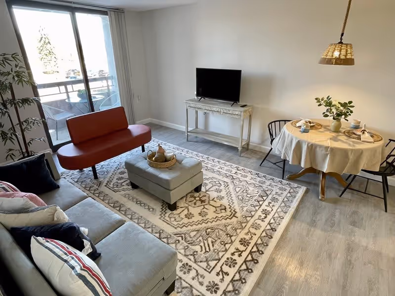 A bright living room with a gray sofa adorned with pillows, a red loveseat, a patterned area rug, and a small ottoman with a decorative tray. A TV sits on a narrow console table against the wall. To the right, there is a round dining table covered with a white tablecloth, set for two with plates, cups, and a vase with greenery. A sliding glass door leads to an outdoor balcony with chairs and a small table.
