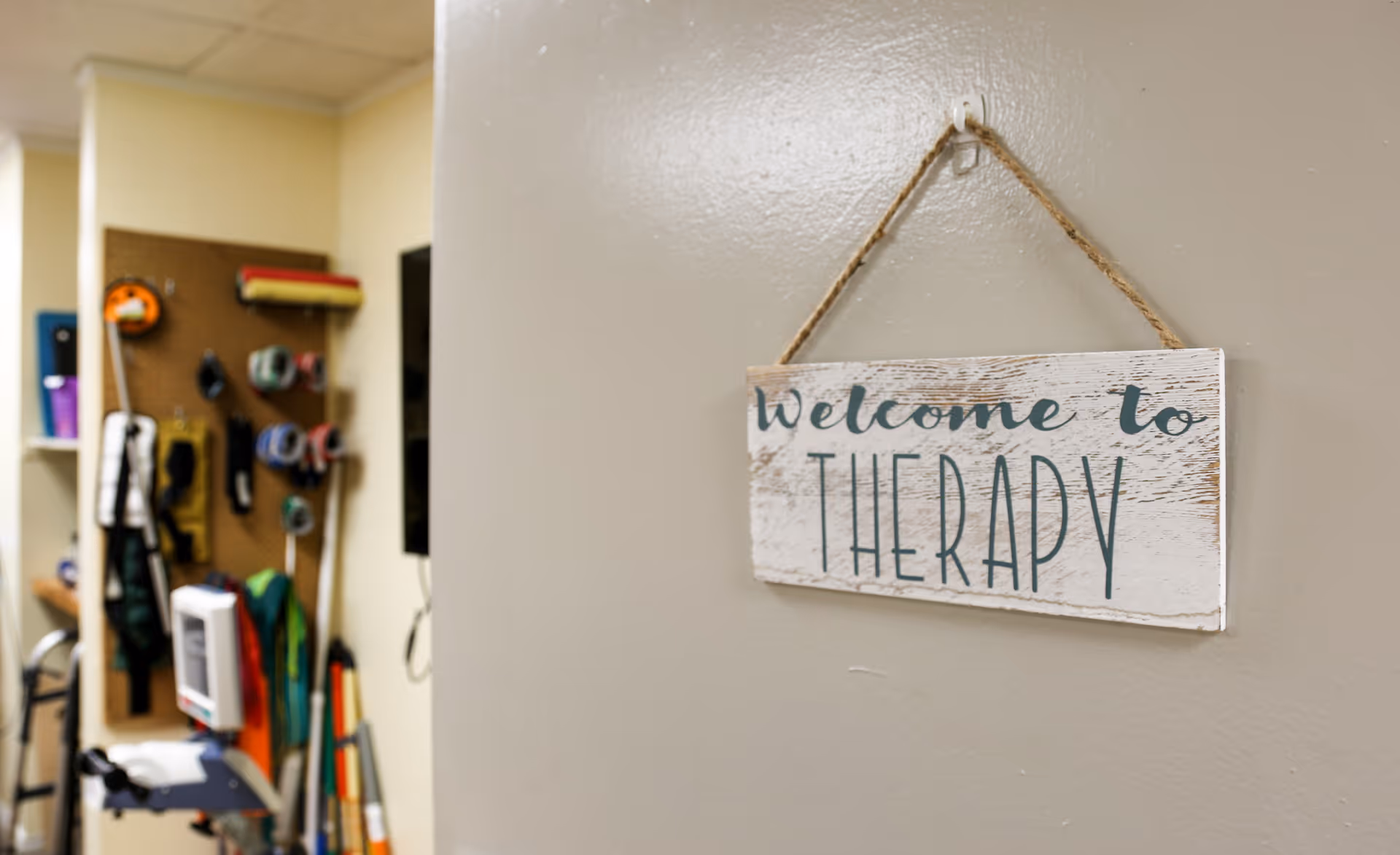 A close-up of a wooden sign hanging on a beige wall that reads 'welcome to THERAPY'. In the background, there is a therapy room with various exercise and rehabilitation equipment, including resistance bands and other physical therapy tools.