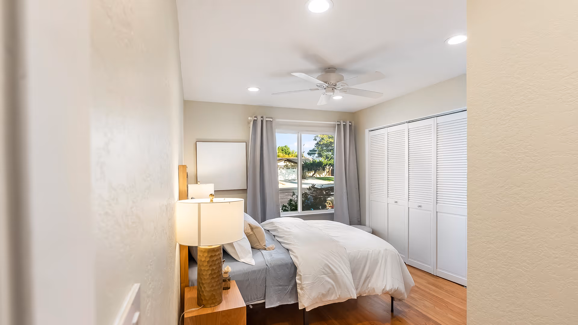 Bright bedroom with a made bed, bedside lamp, ceiling fan, window with curtains, and a wall of white closets.